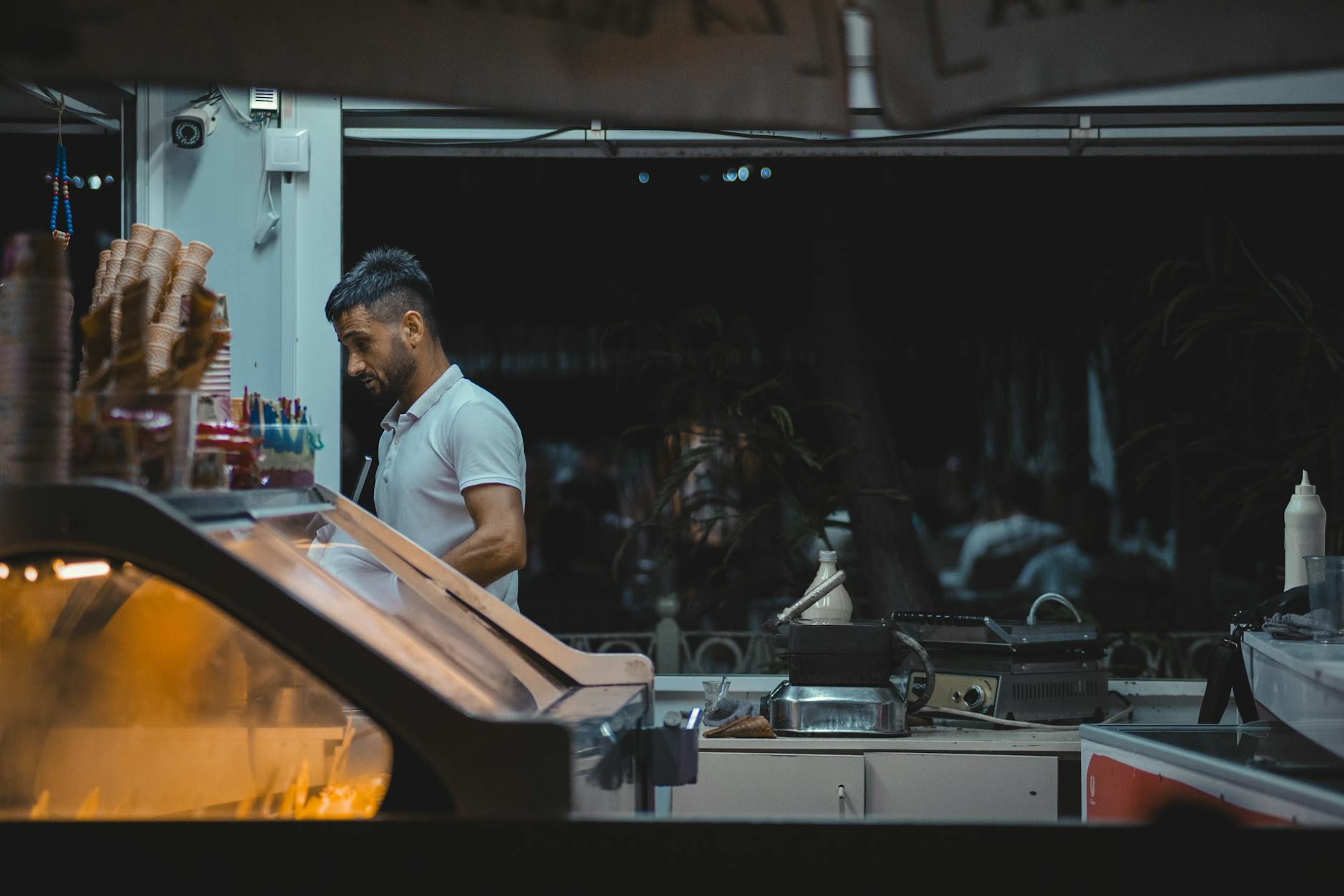 A man working behind the counter of an ice cream shop at night, surrounded by cones and equipment.